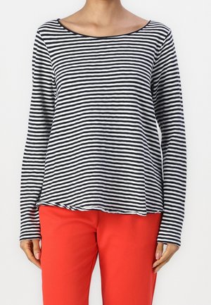 Femme portant un haut à manches longues à rayures horizontales noires et blanches, associé à un pantalon rouge vif, debout devant un fond uni.