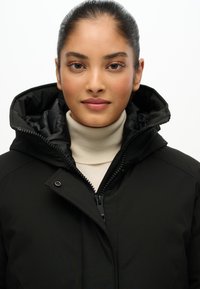 Czarna ocieplana kurtka z wysokim kołnierzem i zapinana na zamek. Posiada kaptur i błyszczące wykończenia. Nosi się ją na kremowym golfie.