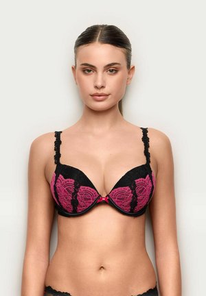 Giovane donna con capelli tirati all'indietro che indossa un reggiseno in pizzo nero e rosa, rivolta verso la parte anteriore su uno sfondo chiaro e uniforme.