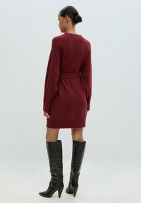 Burgunderfarbendes Strickkleid mit tailliertem Gürtel, langen Ärmeln und geripptem Saum. Kombiniert mit schwarzen, knielangen, hochhackigen Stiefeln mit glatter Textur.