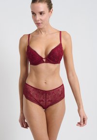 Reggiseno in pizzo bordeaux con scollatura profonda, dotato di spalline regolabili e motivi floreali; culotte in pizzo coordinate con bordi arrotondati.