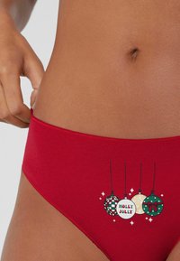 Bikini rojo de algodón con un acabado suave. La parte frontal presenta un diseño festivo con cuatro adornos colgantes, texto "HOLLY JOLLY" y acentos de estrellas.