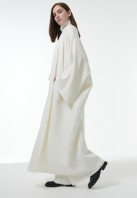 Vrouw in een lange witte coltrui jas en wijde broeken met zwarte schoenen, staande voor een effen witte achtergrond.