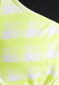 Top desportivo em amarelo neon texturizado, com padrão em colmeia e riscas brancas; tecido preto contrasta na gola.