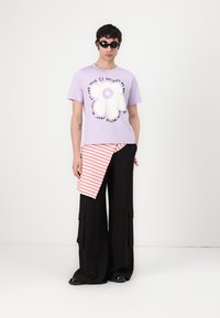 Lila t-shirt med kort ärm och en vit blomma grafik samt svart text, i kombination med svarta vida byxor och ett draperat lager med röda ränder. Svarta skor.