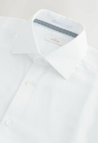 Valkoinen paitapusero, jossa on muotoiltu kaulus ja hienovarainen pintakuvio. Kauluksen sisäosa on sinivalkoinen pilkkukuosisella. Kiinnitetty valkoisilla napeilla.