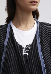 Svart och grå randig cardigan med trasiga blå kanter, lager på en vit t-shirt med en svart blommig grafisk design.