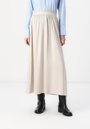 Femme portant une longue jupe beige avec une taille élastique, une chemise rayée bleu et blanc, et des bottes noires mi-mollet, debout sur un sol blanc.