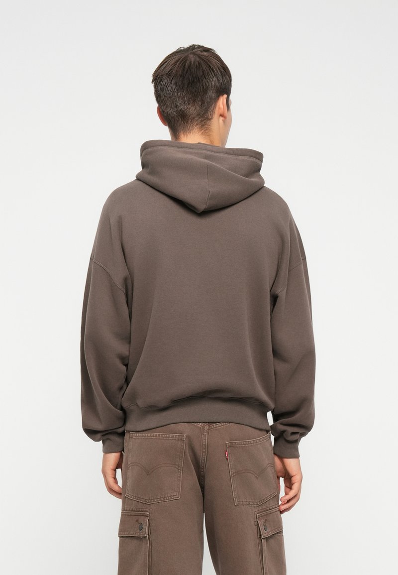 Brązowy hoodie z dużym, oversize'owym kapturem, ściągaczami na rękawach i u dołu; miękka tekstura; noszony z pasującymi brązowymi spodniami cargo.