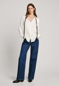 Camicia bianca con bottoni e maniche lunghe, abbinata a jeans a gamba larga blu scuro e stivaletti neri. Tessuto liscio e vestibilità comoda.