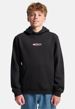 Jongen met krullend rood haar en sproeten, gekleed in een zwarte hoodie met een klein roze logo en een blauwe spijkerbroek, staand tegen een eenvoudige achtergrond.