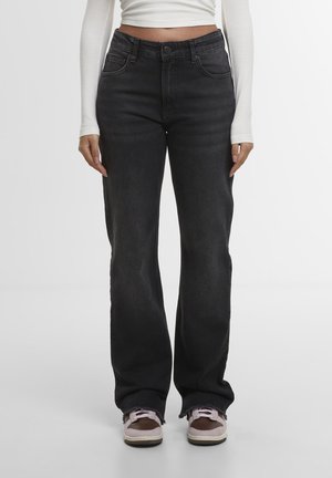 Nő fekete, magasderekú, széles szárú farmert visel, amelynek fodros szegélye van, fehér hosszú ujjú, haspóló, és rózsaszín-barna tornacipőt fehér háttér előtt.