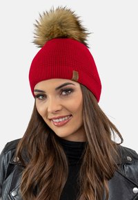 Căciulă tricotată roșie cu un pompon din blană sintetică în vârf, având o textură ribbed și o etichetă mică din piele pe margine.