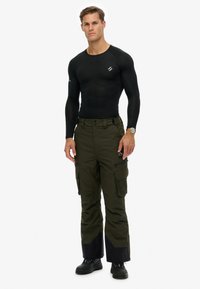 Zwart thermoshirt met lange mouwen en olijfgroene cargobroek met meerdere zakken, voorzien van een zwarte zoom aan de onderkant en zwarte schoenen.