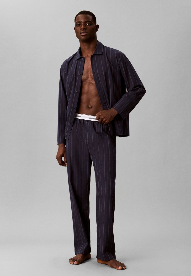 Homme portant un pyjama rayé foncé avec une chemise ouverte, montrant sa poitrine nue et la bande blanche de son sous-vêtement Calvin Klein, debout pieds nus.