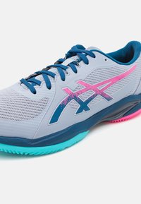 Chaussure de sport grise avec tige en mesh, lacets bleus et accents roses. Elle présente une semelle turquoise et un design texturé pour l'adhérence.