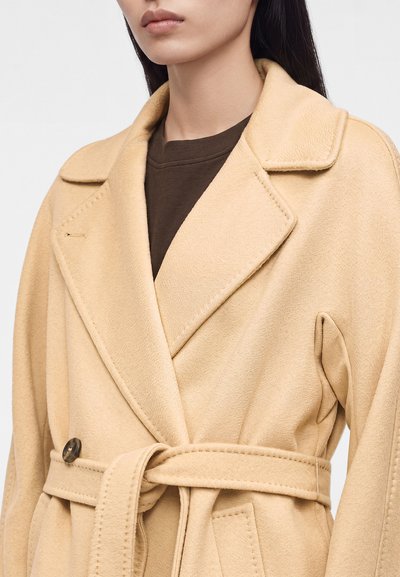 Femme portant un manteau en laine beige ceinturé sur un haut marron foncé avec un large col et des poches avant.