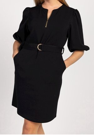 Femme portant une robe noire ceinturée avec des manches bouffantes mi-longues et une fermeture éclair dorée au niveau de l'encolure, mains dans les poches de la robe.