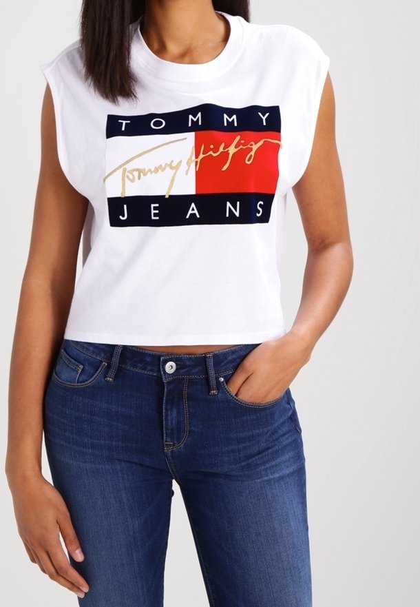 Bílý bezrukávový crop top s grafickým designem "TOMMY JEANS" v červené, námořnické a zlaté barvě. Kombinováno s tmavě modrými džíny.