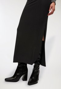 Maxi gonna nera a coste con spacco laterale, abbinata a stivali neri in pelle con tacco alti fino al ginocchio, caratterizzati da punta quadrata e tacco spesso.