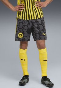 Pantaloni scurți negri și gri cu un model de fagure, logo BVB galben, șosete galbene până la genunchi și un tricou galben cu dungi deschise.