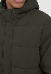 Mann, der eine dunkelgrüne, gepolsterte Winterjacke mit hohem Kragen und Kapuze trägt, von Kinn bis Brust gezeigt.