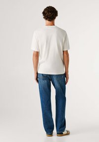 Man die met de rug naar de camera staat, gekleed in een wit shirt met korte mouwen, blauwe jeans en witte sneakers op een effen achtergrond.