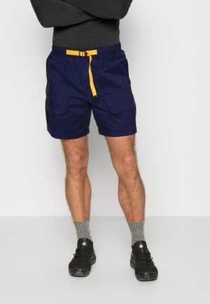 Granatowe spodenki z żółtym sznurkiem, dwoma bocznymi kieszeniami i luźnym krojem. Noszone z szarymi skarpetkami i czarnymi butami.