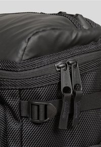 Musta kangaskassi, joka sisältää tekstuuroitua materiaalia, kaksinkertaiset vetoketjut ja pehmustetun yläosan. Mukana on solkistrappi turvalliseen sulkemiseen.