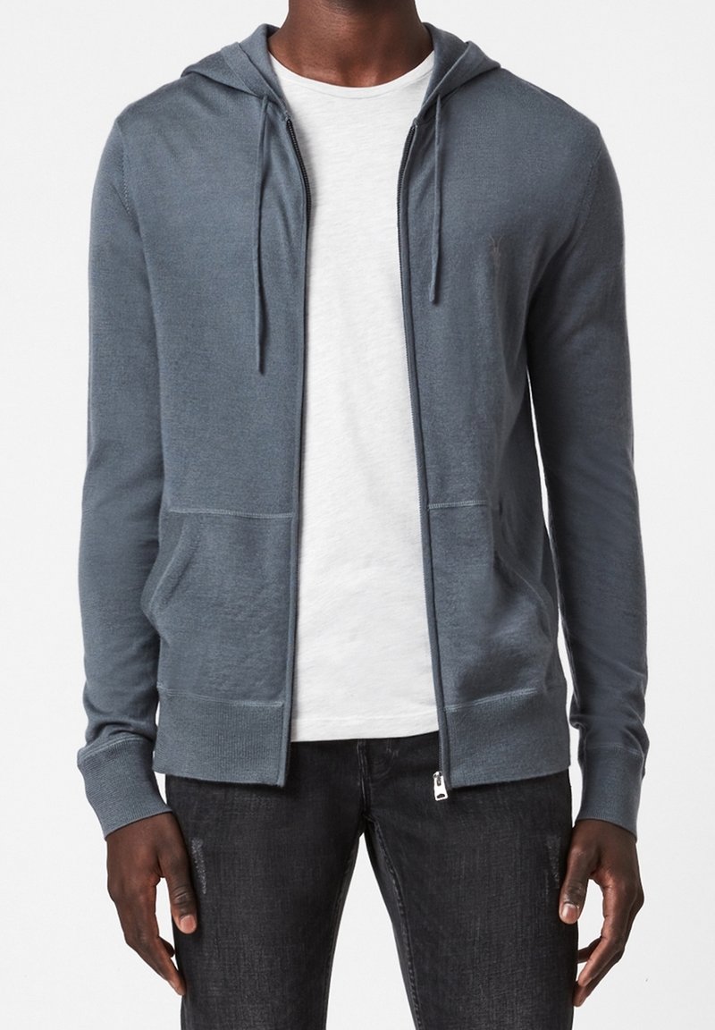 Haut à capuchon gris zippé en tissu doux, avec une capuche, des poches avant et des poignets côtelés, superposé sur une chemise blanche.