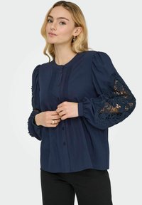 Blusa blu navy con maniche a sbuffo, dettagli in pizzo sui polsini, chiusura con bottoni e accenti plissettati. Il tessuto è liscio e leggero.