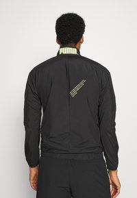 Svart lättviktsjacka med hög krage och en grön logotyp. Innehåller en bakre söm och tryckta textdetaljer. Elastiska ärmslut och nederkant.