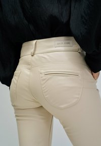 Pantalons beiges ajustés avec des poches arrière et une étiquette en cuir portant la mention "Salsa Jeans", portés avec un haut noir, mettant l'accent sur la taille et les hanches.