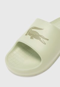 Ljusgrön slide-sandal i syntetmaterial, med öppen tå, texturerad fotbädd och en grå krokodillogga på remmen.
