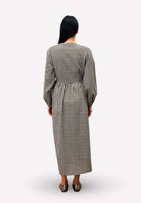 Femme aux longs cheveux noirs portant une robe midi à manches longues à carreaux noirs et blancs et des chaussures bronze, se tenant de dos sur un fond blanc.