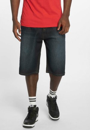 Shorts en denim bleu foncé avec des coutures orange, coupe au genou, portés avec des baskets noires et des chaussettes rayées blanches, associés à une chemise rouge.