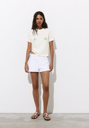 Femme debout contre un mur blanc, portant un t-shirt crème avec des fleurs bleues, un short blanc et des sandales noires avec des coquillages blancs.