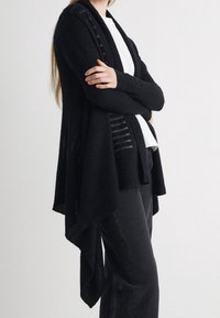 Svart stickad cardigan med öppen front, långa ärmar och asymmetrisk fåll. Har genomskinliga randiga paneler längs de övre sidorna.