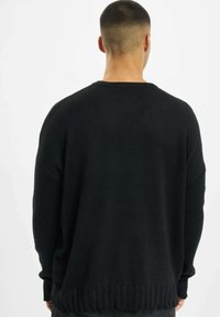 Pull en maille noire avec une coupe décontractée, texture côtelée, col rond et détails d'ourlet effiloché. La vue arrière montre des manches amples et un design simple.