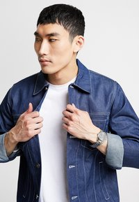 Veste en denim, bleu foncé avec des coutures blanches, présente une fermeture à boutons à l'avant et des poignets bleu clair. Portée par-dessus un t-shirt blanc avec une montre en argent.
