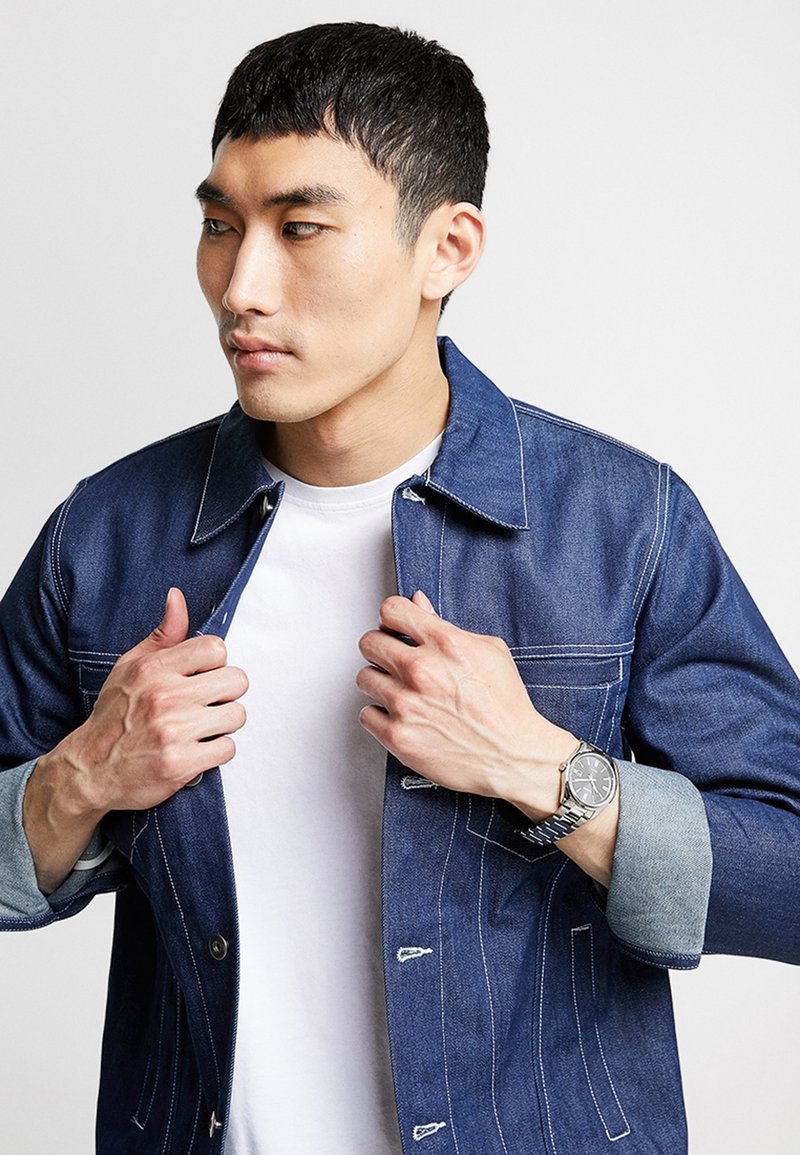 Veste en denim, bleu foncé avec des coutures blanches, présente une fermeture à boutons à l'avant et des poignets bleu clair. Portée par-dessus un t-shirt blanc avec une montre en argent.