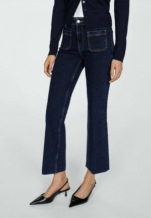 Granatowe dżinsy dzwony o wysokim stanie, z kontrastującym przeszyciem, tylnymi kieszeniami i gładkim wykończeniem. Połączone z czarnymi szpilkami z ostrym noskiem.