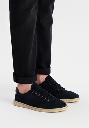 Mørkeblå suede sneakers med lavt design, rund tå og sorte snørebånd. Cremefarvet gummisål. Rullede sorte jeans synlige ovenfor.