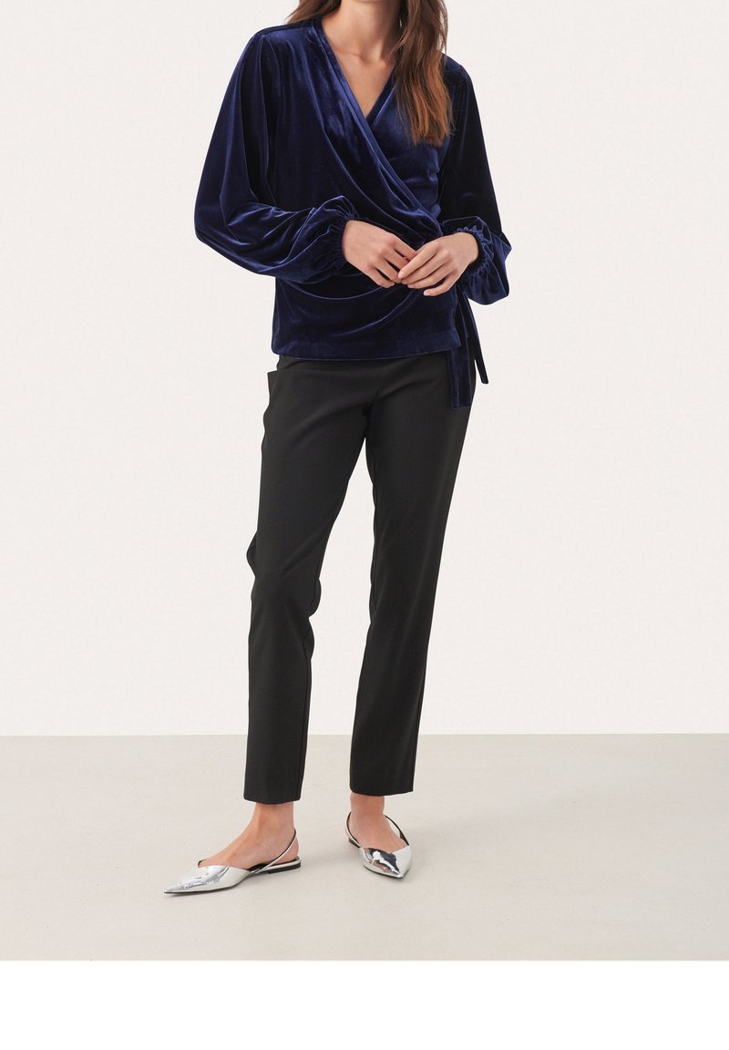 Femme portant un chemisier cache-cœur en velours bleu foncé, un pantalon noir ajusté et des chaussures plates argentées à bout pointu, debout sur un fond uni.