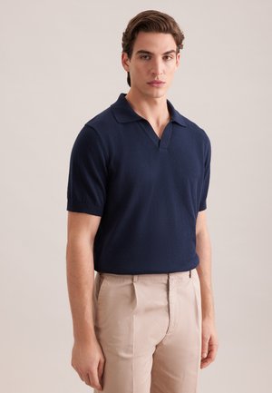 Jeune homme portant un polo bleu marine à manches courtes et un pantalon beige, debout devant un fond clair uni.