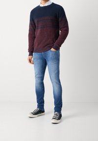 Hombre vestido con un suéter a rayas rojas y azul marino, jeans ajustados de color azul claro y zapatillas negras de caña baja con cordones blancos, de pie frente a un fondo blanco.