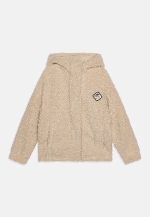 Hoodie en fausse fourrure beige avec une fermeture éclair frontale, deux poches latérales et un patch étiquette tissée présentant une forme de diamant et du texte. Texture douce.