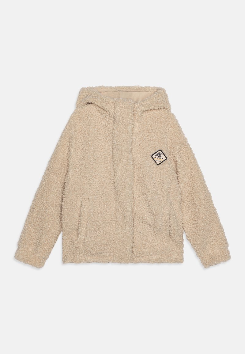 Hoodie en fausse fourrure beige avec une fermeture éclair frontale, deux poches latérales et un patch étiquette tissée présentant une forme de diamant et du texte. Texture douce.