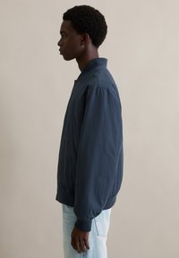 Granatowa bomberka o gładkiej teksturze, zamek błyskawiczny z przodu, elastyczne mankiety i boczne kieszenie, zestawiona z jasnoniebieskimi dżinsami. Luźny fason.