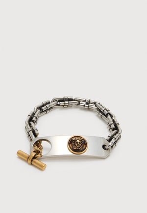 Brățară cu lanț din argint cu o placă dreptunghiulară cu un emblema aurită cu capul Meduzei și o închizătoare aurie tip toggle, pe fundal alb.